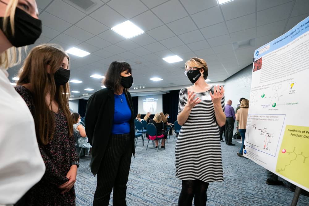 Scholar wearing mask presenting poster to three guests wearing masks.